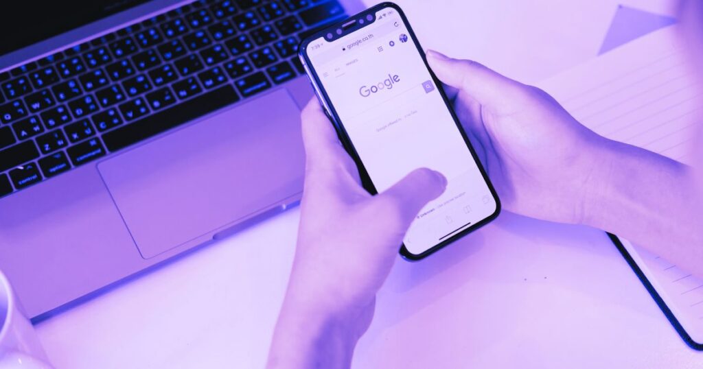 Deux mains tiennent un smartphone avec la page de Google, avec un clavier d'ordinateur dans le fond