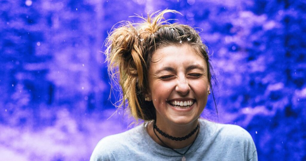 Une femme ferme les yeux en riant. Elle savoure la pluie qui tombe sur un chemin en forêt, symbolisant la sérénité de certaines personnes cultivées