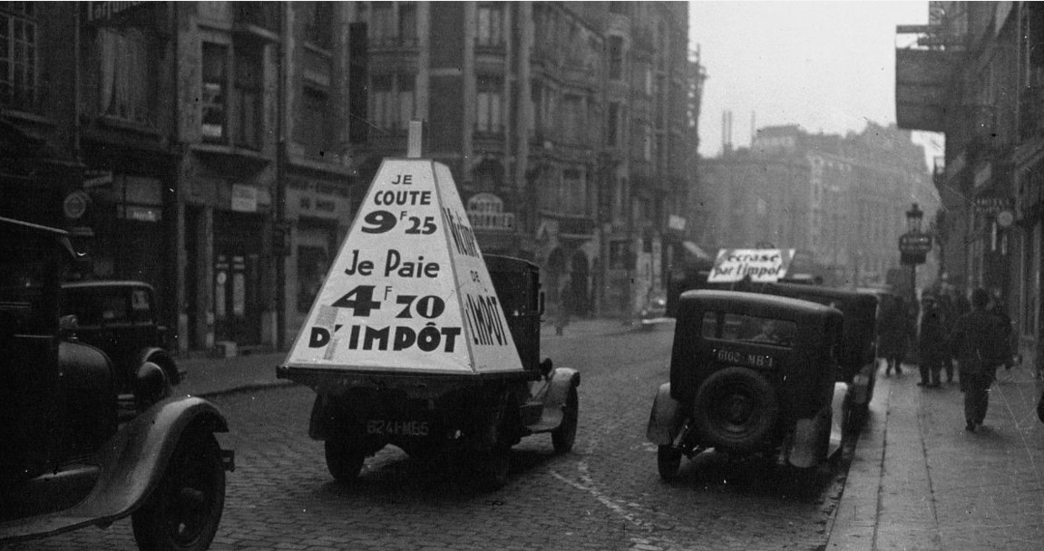 Photographie en noir et blanc d'une rue parisienne dans les années 1930, avec un véhicule portant une pancarte de protestation contre les impôts, symbole de la crise économique de la Grande Dépression