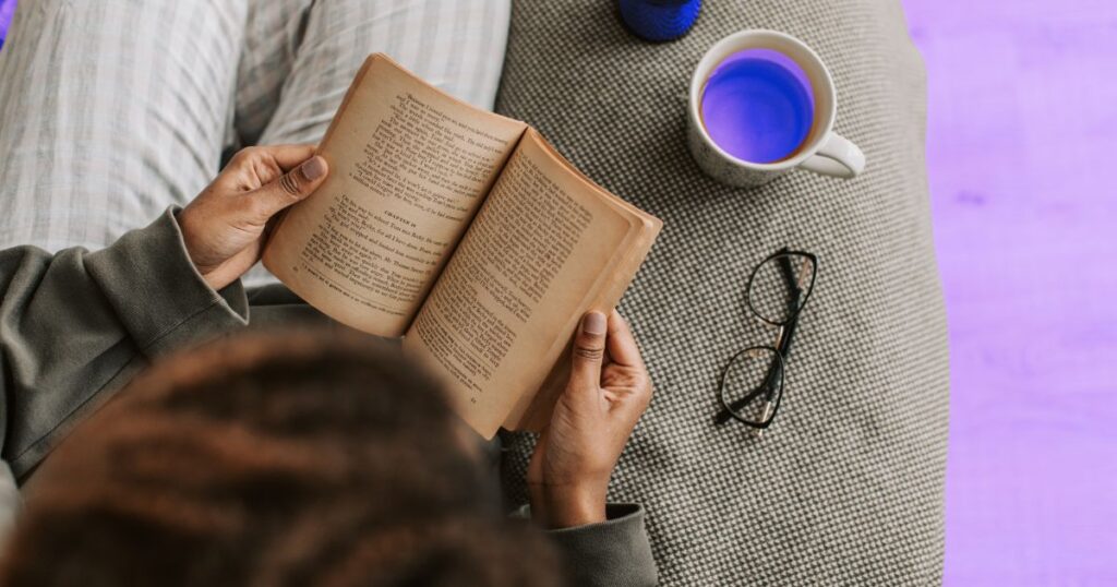 Vue plongeante d'une femme lisant un livre sur un canapé, avec une tasse de café et une paire de lunettes posées près d'elle