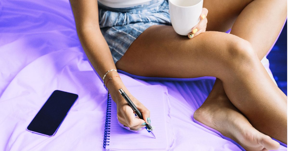 Une femme prend des notes, assise sur son lit, avec une tasse de thé à la main